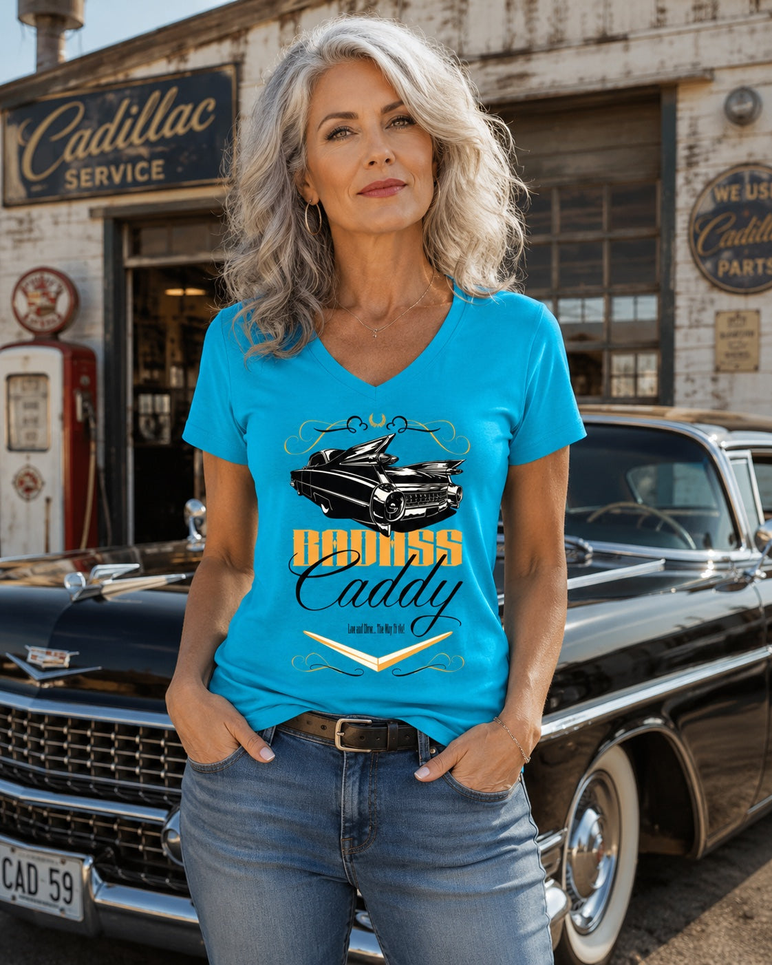 A woman with gray hair stands in front of a vintage black  Cadillac and Service shop, wearing a bright blue V-neck graphic t-shirt with a black Cadillac car illustration and yellow text reading 'Badass Caddy'.