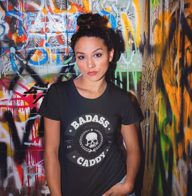 Woman wearing a black t-shirt with 'BADASS CADDY' text in front of a graffiti-covered wall.